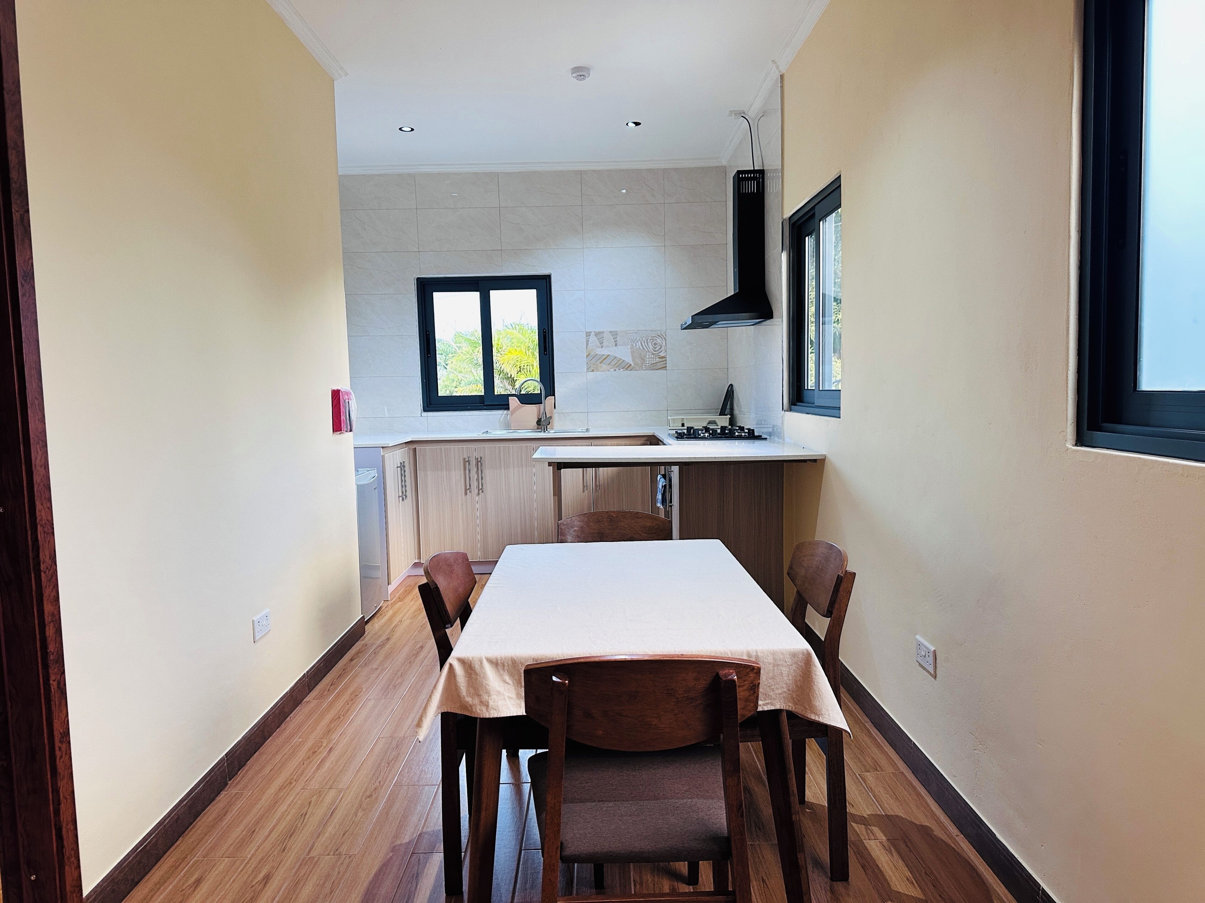 A narrow hallway leading to a kitchen with a dining table and chairs, a window, and a tiled backsplash.