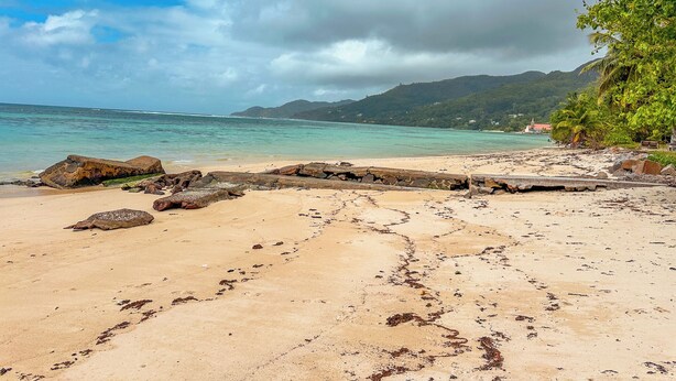 Una playa con agua cristalina, formaciones rocosas y una cadena montañosa al fondo.