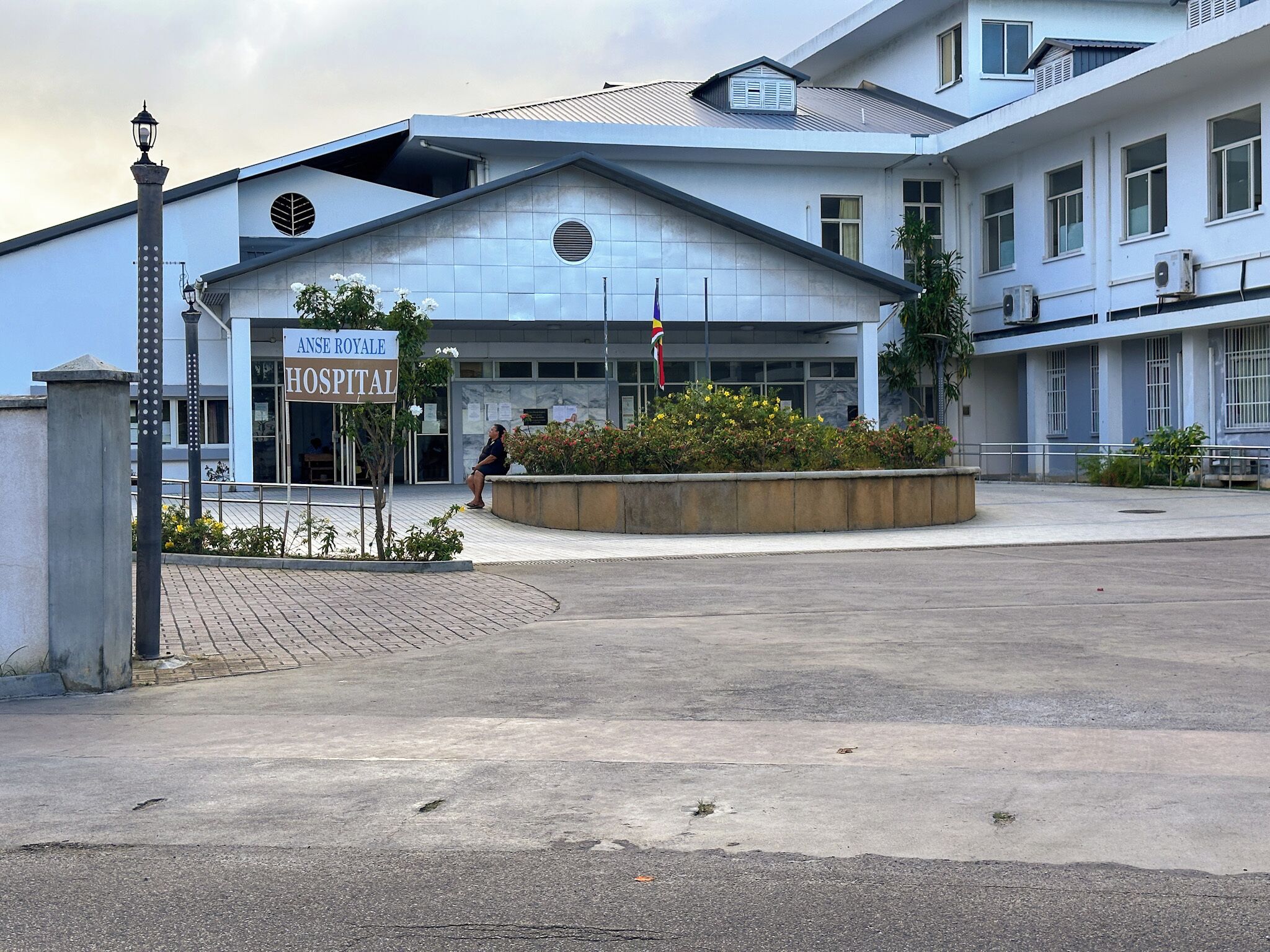 A hospital building with a sign reading 'ANSE ROYALE HOSPITAL,' a paved entrance, and a flagpole with a flag.