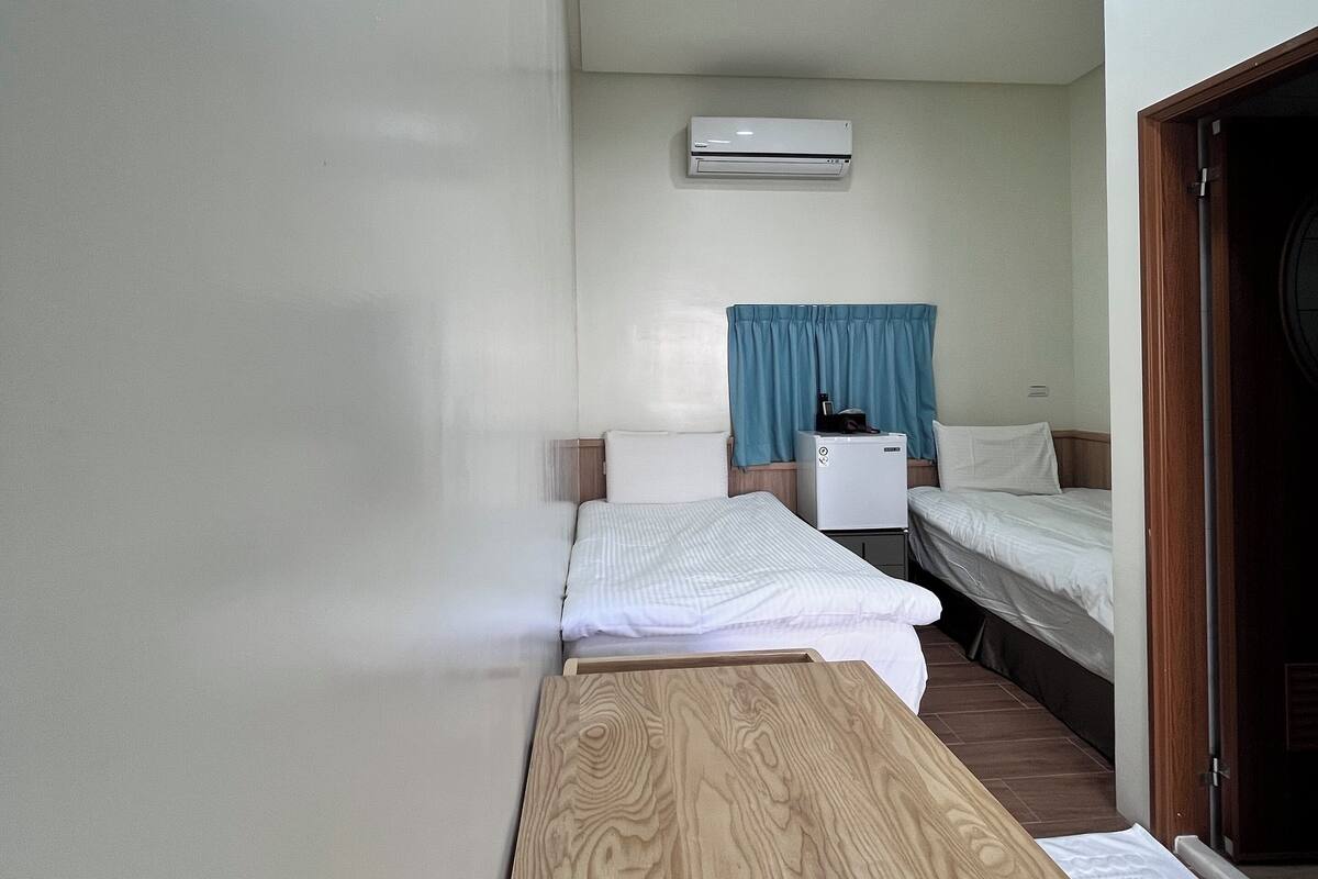 Una habitación pequeña y limpia con una mesa de madera, dos camas, un equipo de aire acondicionado blanco y una cortina azul.