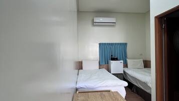 A small, clean room with a wooden table, two beds, a white air conditioning unit, and a blue curtain.