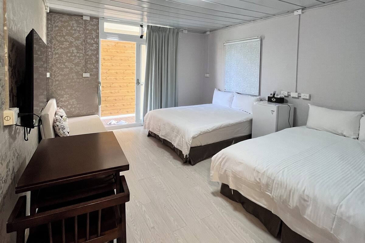 Habitación de hotel con dos camas, una mesa de madera, un televisor y un ventanal con cortinas.