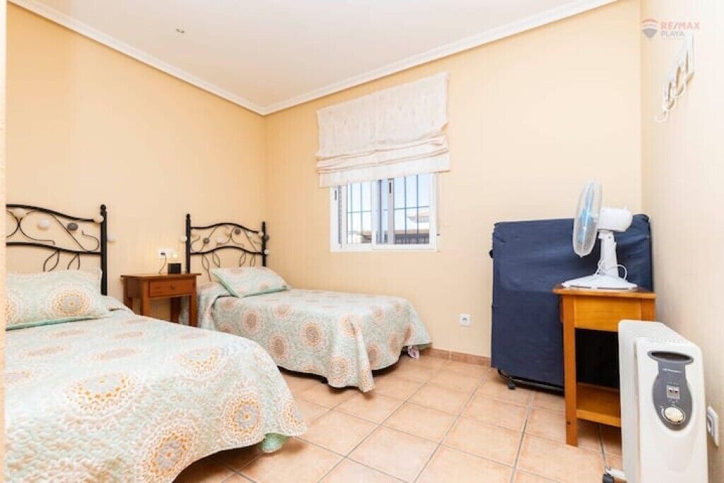 A bedroom with two beds, a window with a white blind, a heater, and a small table with a lamp.