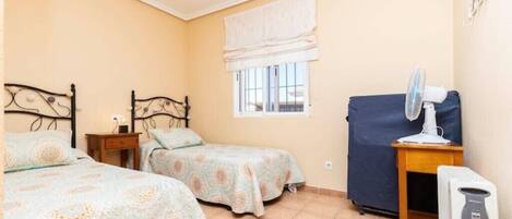 A bedroom with two beds, a window with a white blind, a heater, and a small table with a lamp.
