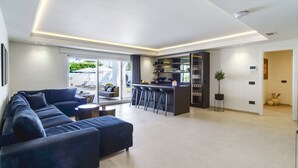 A modern living room with a dark-colored sofa, a small round table, and a kitchen area with a bar and stools. - Luxury Family Villa for in Marbella (Marbella)