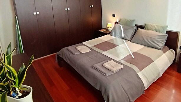 A bedroom with a large bed, a wardrobe, a bedside table with a lamp, and a potted plant. - Dietro la piazza (Marostica)