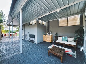 An outdoor patio area with a wooden bench, a sofa, and a table. There is a fire pit and a wooden cabinet with decorative items. - Spacious 4BR Home with Swimming Pool | Free Parking (Picnic Point)