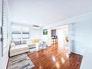 A modern living room with a wooden floor, a sofa, a coffee table, and a dining area with bar stools. - Spacious 4BR Home with Swimming Pool | Free Parking (Picnic Point)