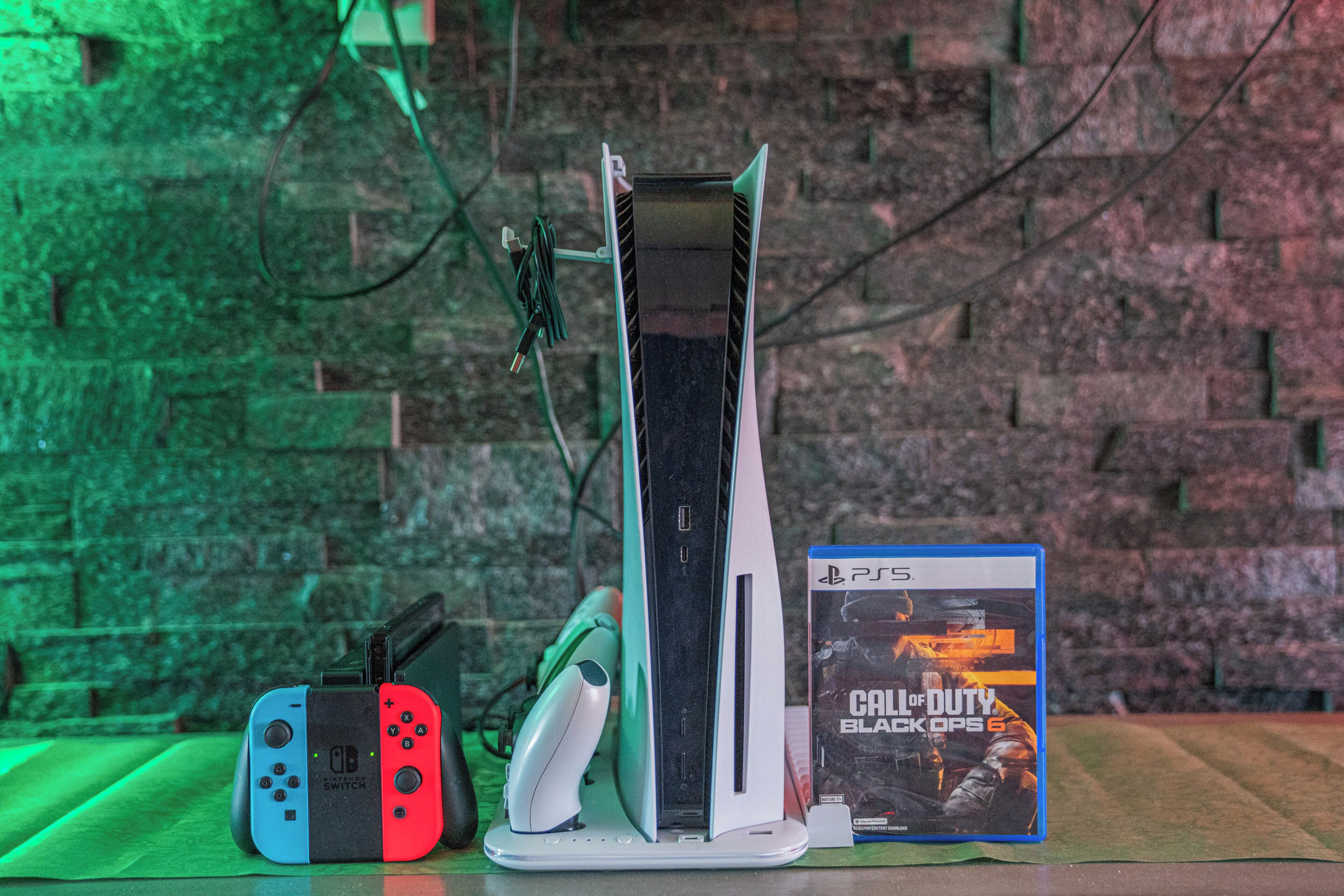 A PlayStation 5 console, two controllers, and a game case on a table with a brick wall background.