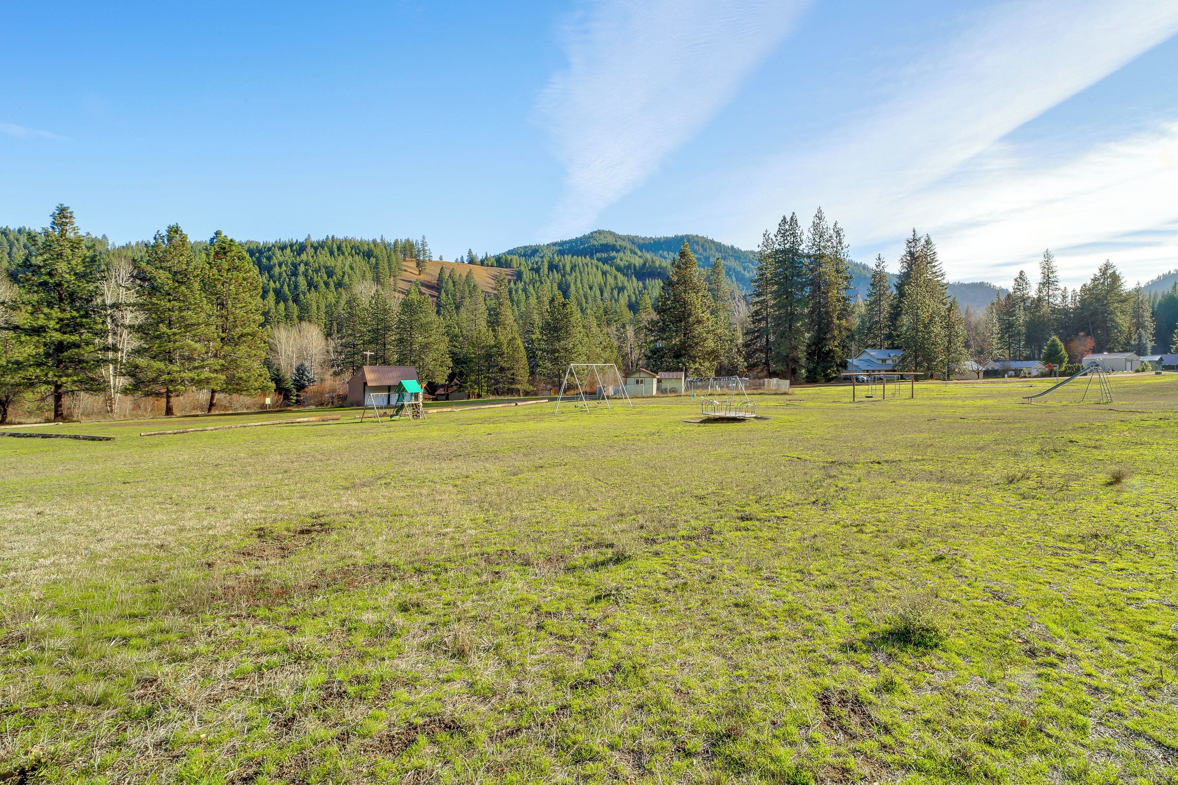 A grassy field with a swing set, a small building, and a forested hill in the background.