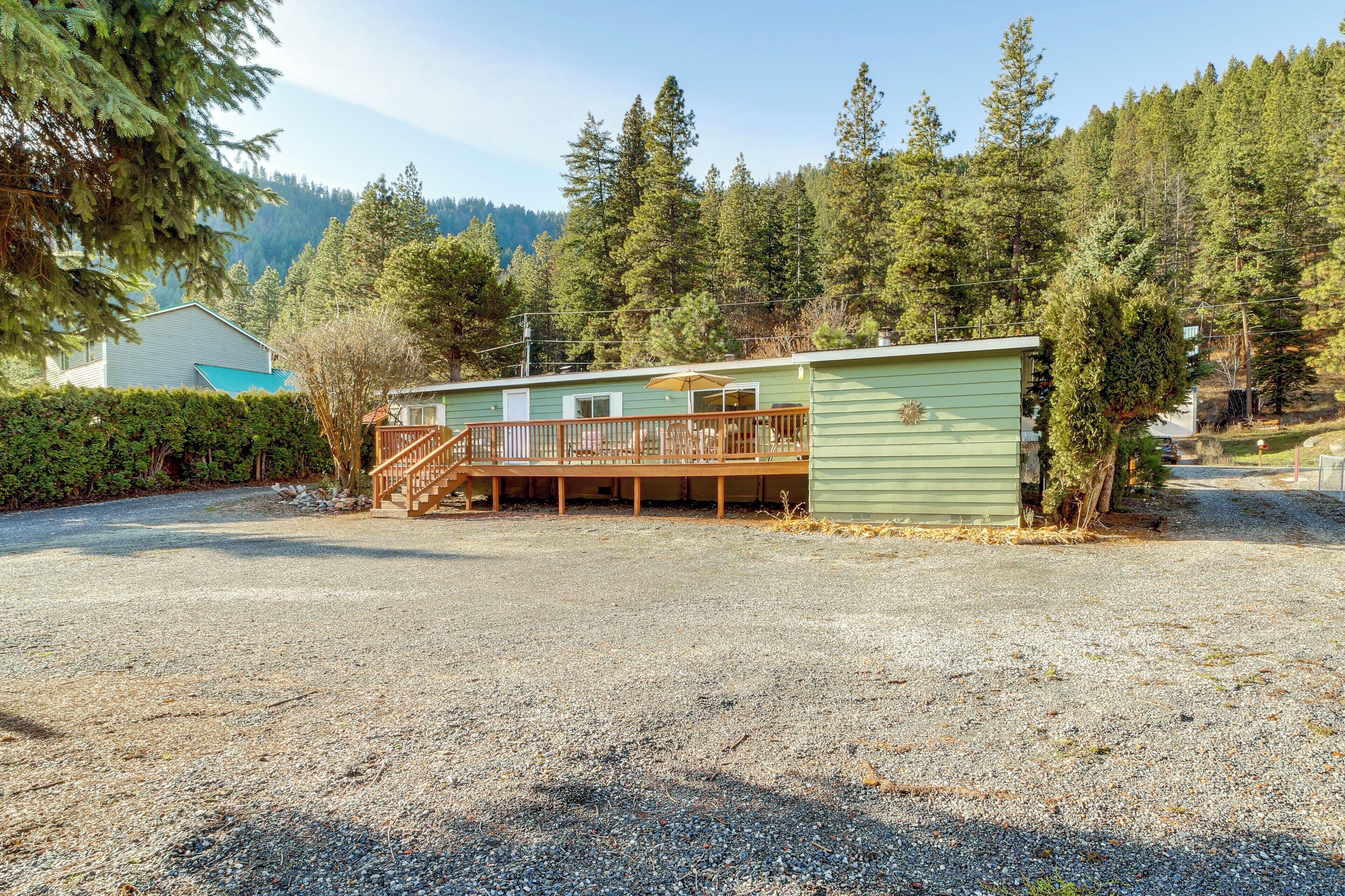A single-story cabin with a wooden deck and a covered porch, surrounded by dense forest and a clear sky.