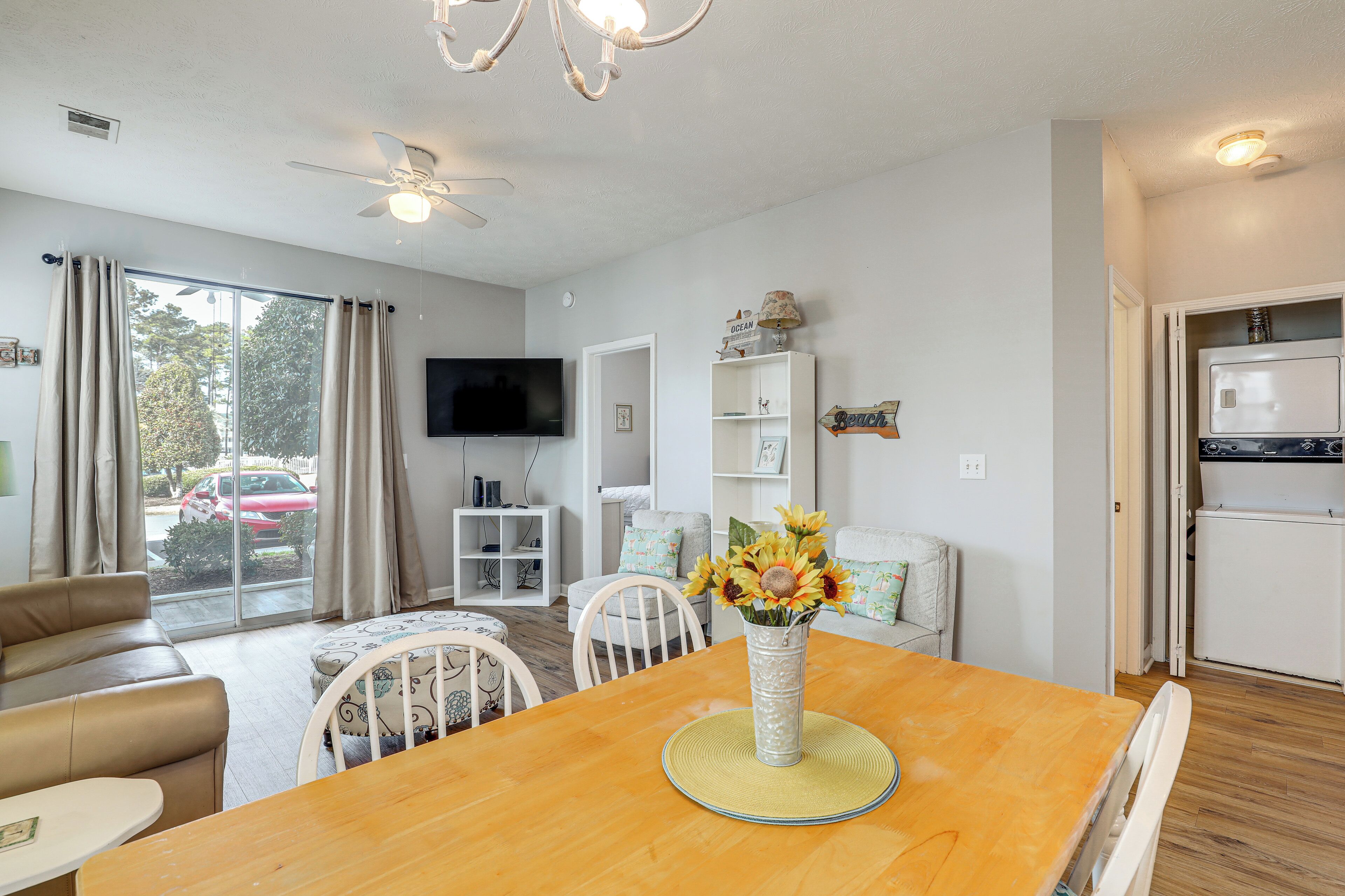 A dining area with a wooden table, white chairs, a vase with flowers, a ceiling fan, and a television in the background.