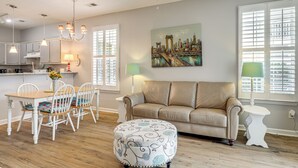 Een woonkamer met een beige bank, een eettafel met stoelen, een rond voetenbankje, en een schilderij van een stadsgezicht.