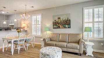 A living room with a beige sofa, a dining table with chairs, a round ottoman, and a painting of a cityscape.