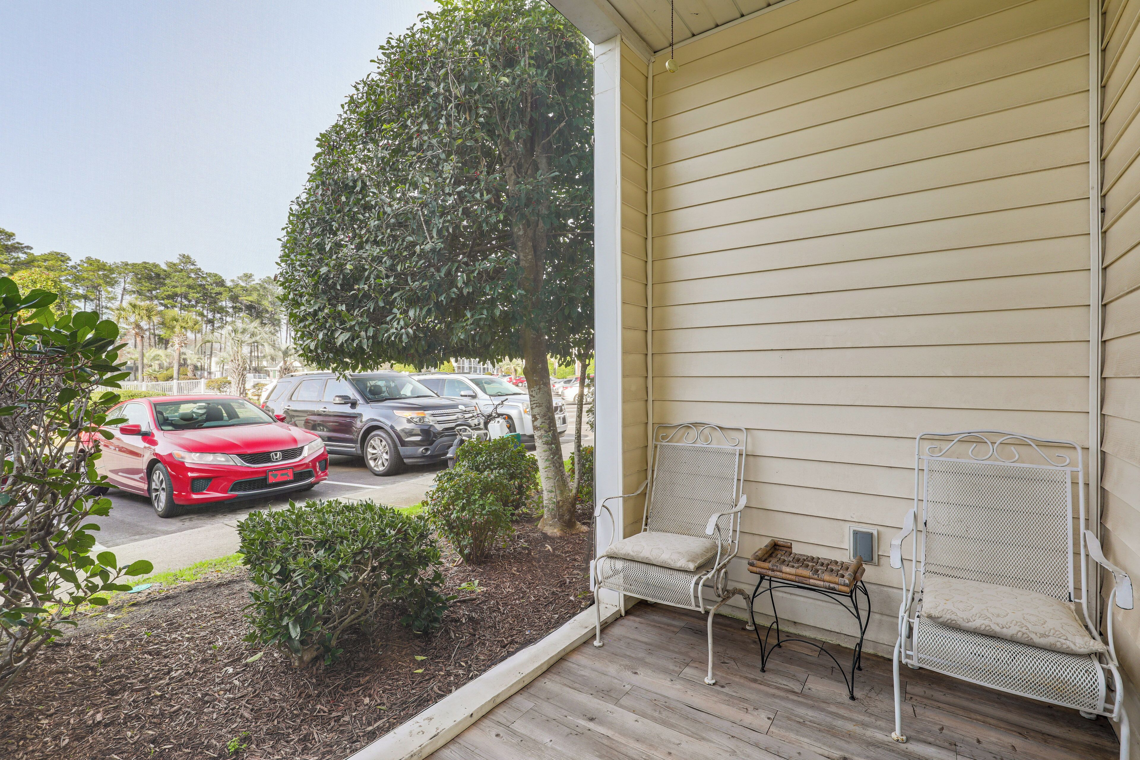 A porch with two chairs and a small table, a red car parked in the background, and a tree.