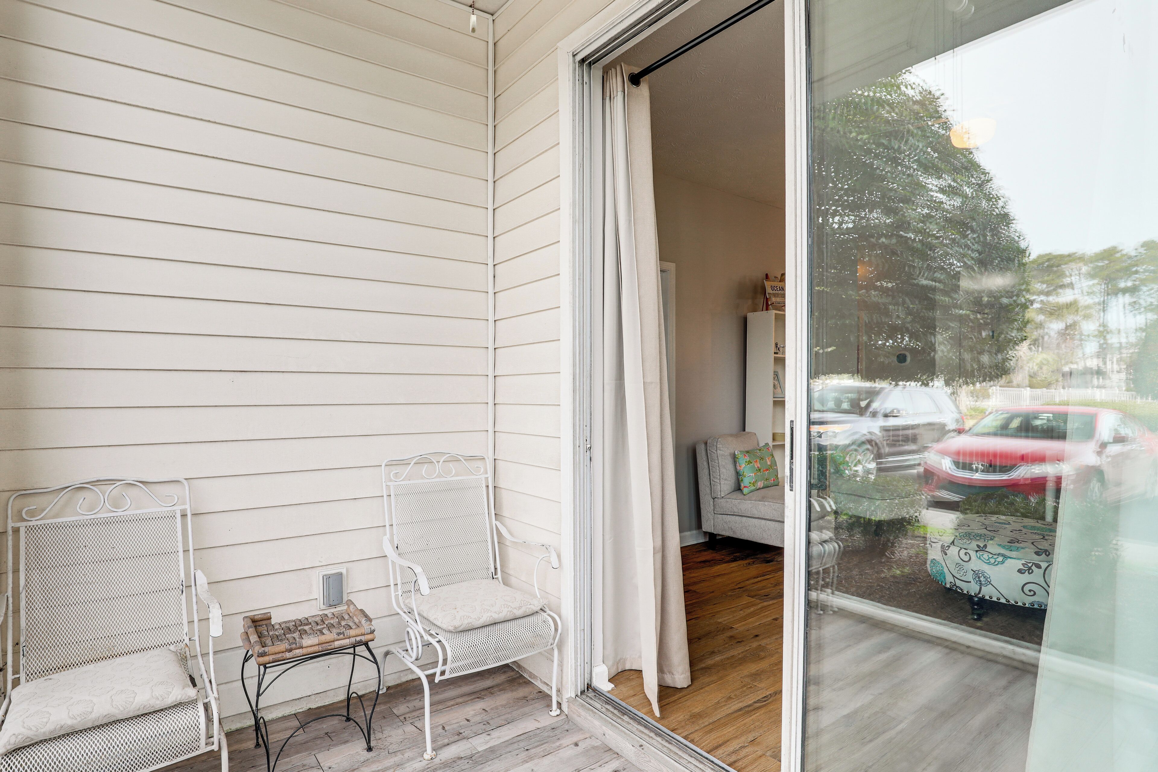 A balcony with two white metal chairs and a small table, a sliding glass door leading to a room with a sofa and a patterned tablecloth.