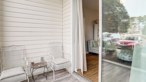 Een balkon met twee witte metalen stoelen en een kleine tafel, een schuifdeur die leidt naar een kamer met een bank en een tafelkleed met een patroon.