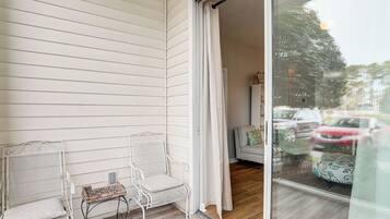 A balcony with two white metal chairs and a small table, a sliding glass door leading to a room with a sofa and a patterned tablecloth.