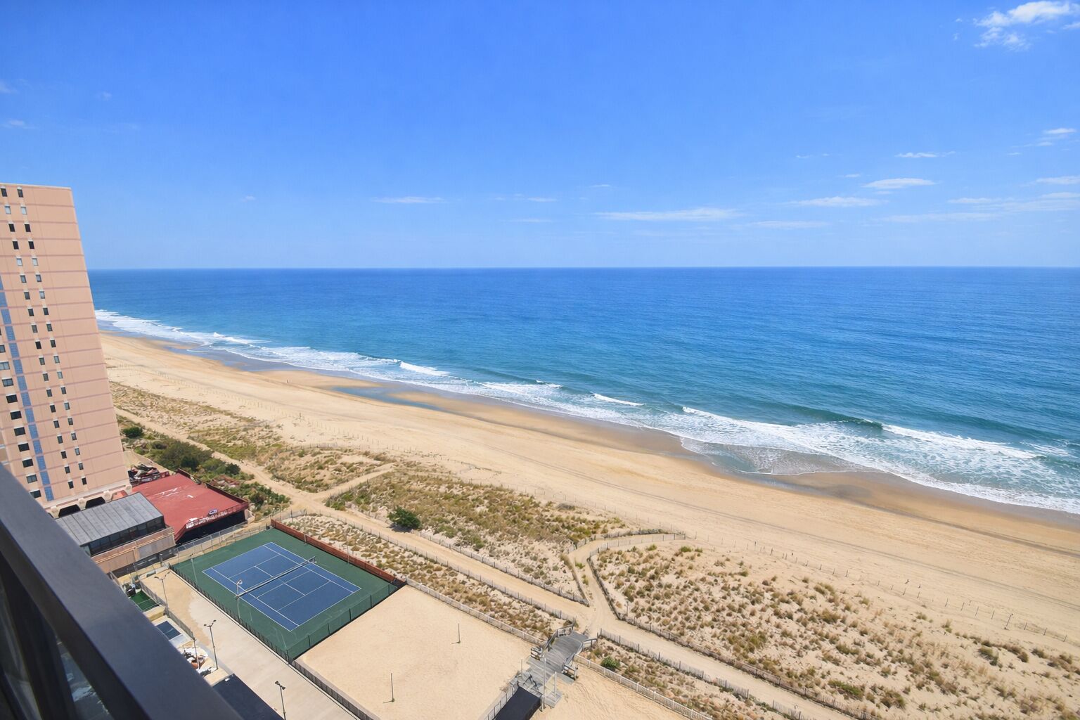 Uitzicht op het strand vanaf een hoog gebouw, met een tennisbaan en een parkeerplaats.