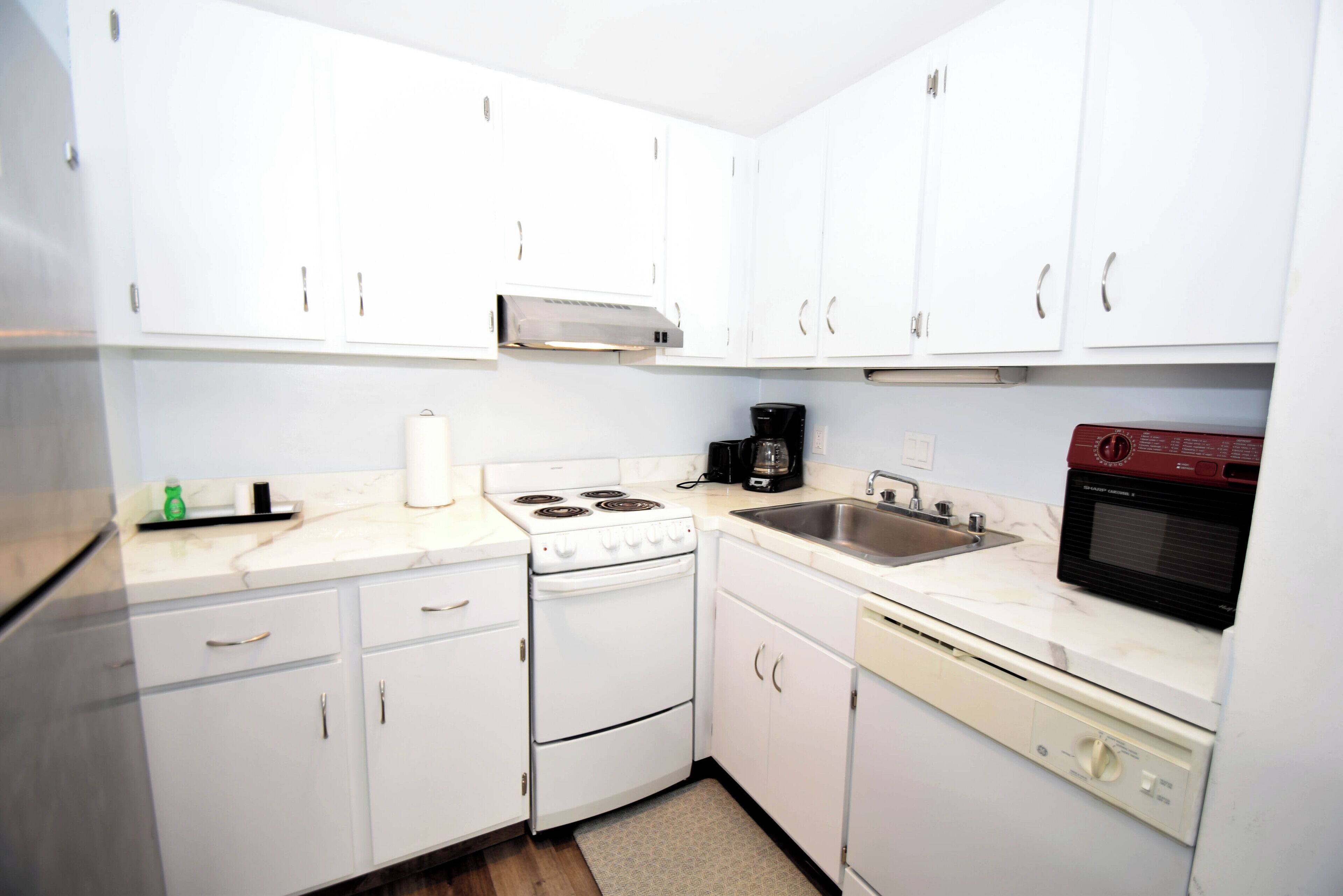 A compact kitchen with white cabinets, a stove, a microwave, a coffee maker, and a dishwasher.