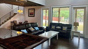 Ein großzügiges Wohnzimmer mit einer schwarzen Ledersofa, einem weißen Couchtisch und einem Essbereich mit Stühlen und Tisch. Eine Treppe mit Geländer, ein Deckenventilator und große Fenster mit Blick ins Freie sind ebenfalls vorhanden.