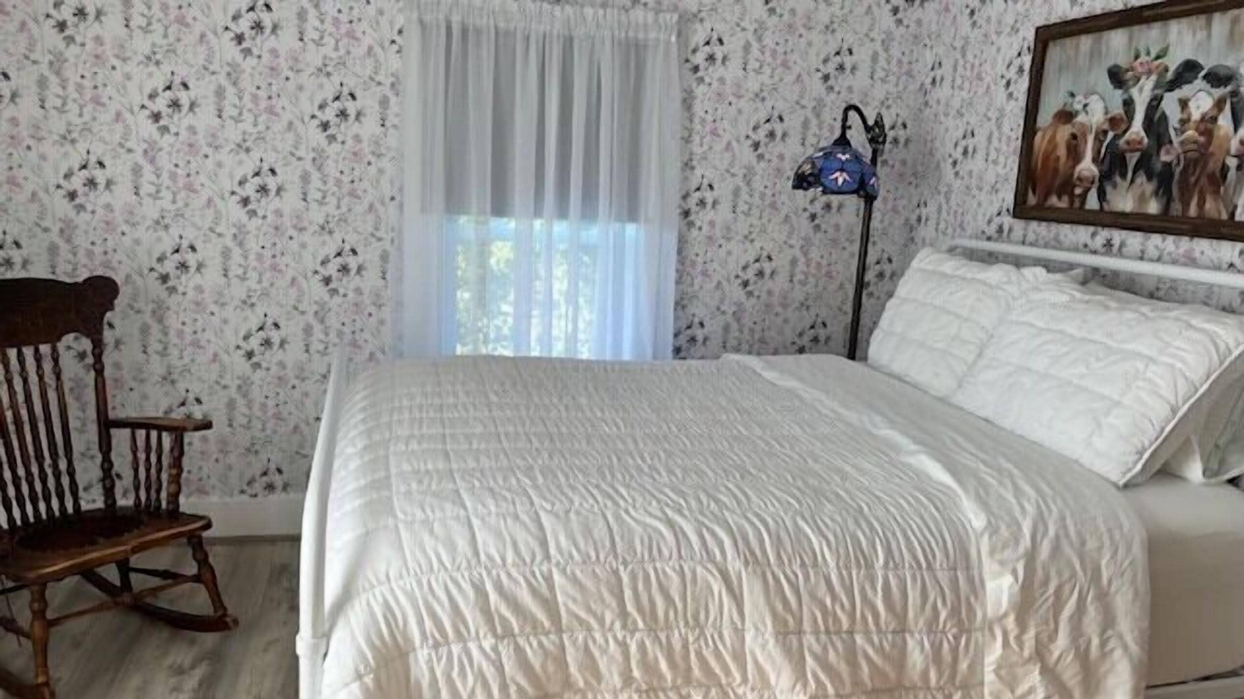 A bedroom with a bed, a rocking chair, a window with sheer curtains, and a wall-mounted lamp.