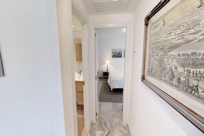 Ein Flur, der zu einem Schlafzimmer mit einem Bett, einem Nachttisch und einer Lampe führt. An der Wand hängt ein großes, gerahmtes historisches Gemälde.