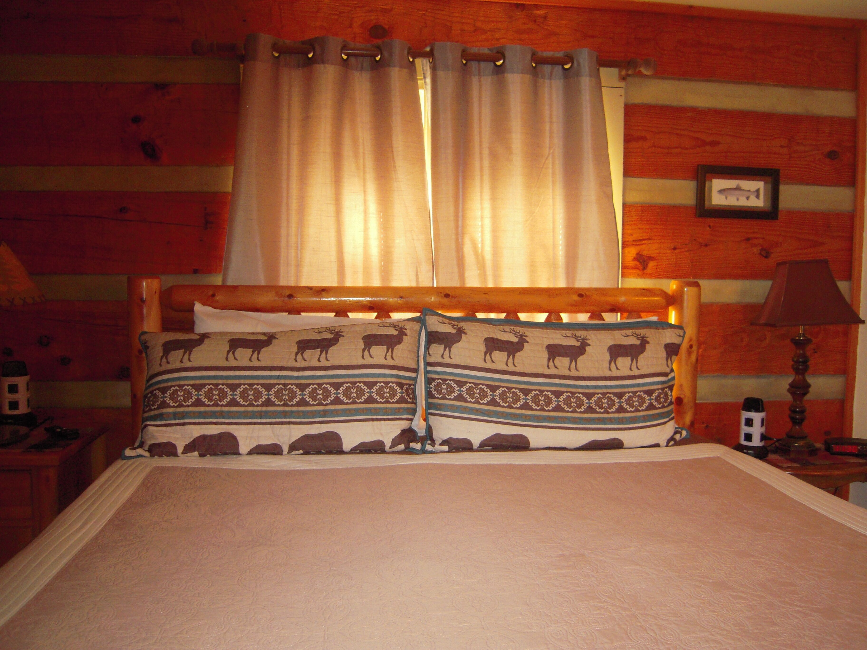Habitació de tipus cabana de fusta amb un llit, cortines, una làmpada i un quadre al mur.