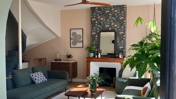 Un salon avec un canapé, une table basse et une cheminée. Un escalier se trouve à gauche et une grande plante à droite.