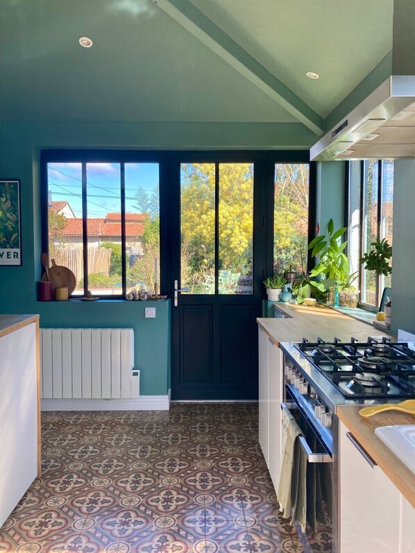 A kitchen with a black door, a window with a view of a house and trees, a gas stove, and a radiator. - A Toulouse woman of character (Lavernose-Lacasse)