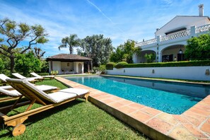 Piscina com espreguiçadeiras, um pequeno gazebo e uma casa branca com cerca branca e vegetação.