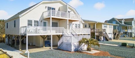 A multi-story beachfront property with a balcony, a small garden, and a clear sky.