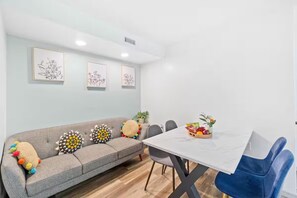 Una sala de estar moderna con un sofá gris, cojines coloridos, una mesa blanca con fruteros y reproducciones botánicas enmarcadas en la pared.