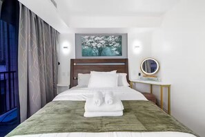 Una habitación de hotel con una cama bien hecha, un cuadro de un árbol, una mesita de noche con espejo y una ventana con cortinas.
