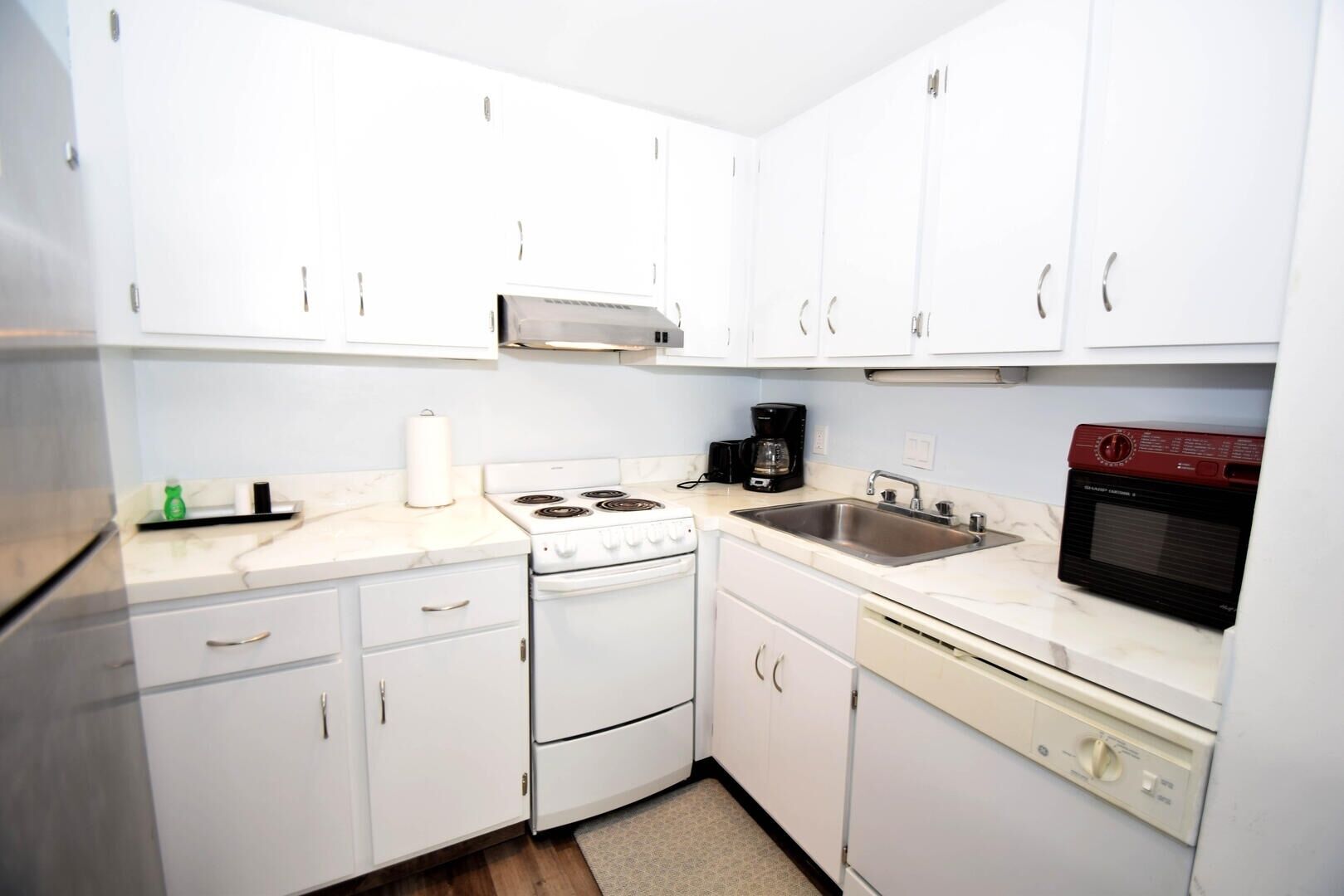 A compact kitchen with white cabinets, a stove, a sink, a coffee maker, and a microwave.
