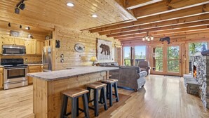 A wooden cabin interior with a kitchen, bar area, seating, and a stone fireplace. - A View To A Dream 4 Bedroom Home (Sevierville)