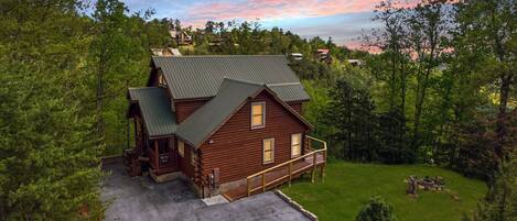 A wooden cabin with a porch, a fenced yard, and a view of the surrounding forest.