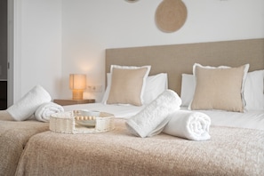 Una cama bien hecha con ropa de cama blanca y beige, una lámpara de noche y una cesta tejida con bolas de madera.