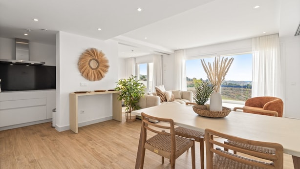 A modern kitchen with white cabinets, a black countertop, and a built-in stove. There is a dining area with a white table and wicker chairs. A large window offers a view of the outdoors. - Alcazaba Lagoon Retreat for 4 (Casares)