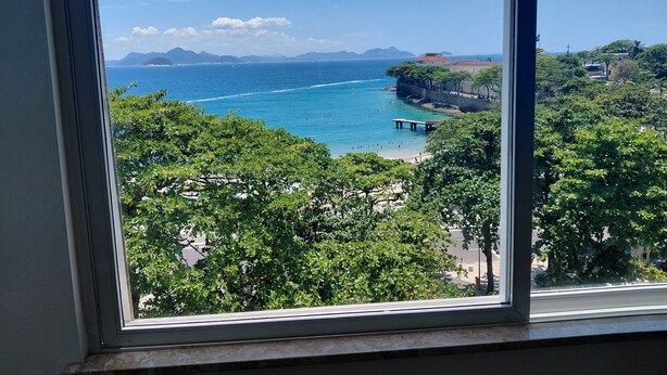 A coastal view through a window, featuring a beach, clear blue sea, distant mountains, and lush greenery. - Atlantic Avenue, Post 6, Incredible View  (Rio de Janeiro)
