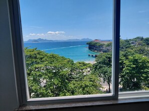 A coastal view through a window, featuring a beach, clear blue sea, distant mountains, and lush greenery. - Atlantic Avenue, Post 6, Incredible View   (Rio de Janeiro)