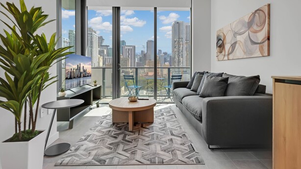 A modern living room with a sofa, a coffee table, and a view of a city skyline through large windows. - 501 First Luxury Corner Studio Rooftop Pool & GYM 1101A (Miami)
