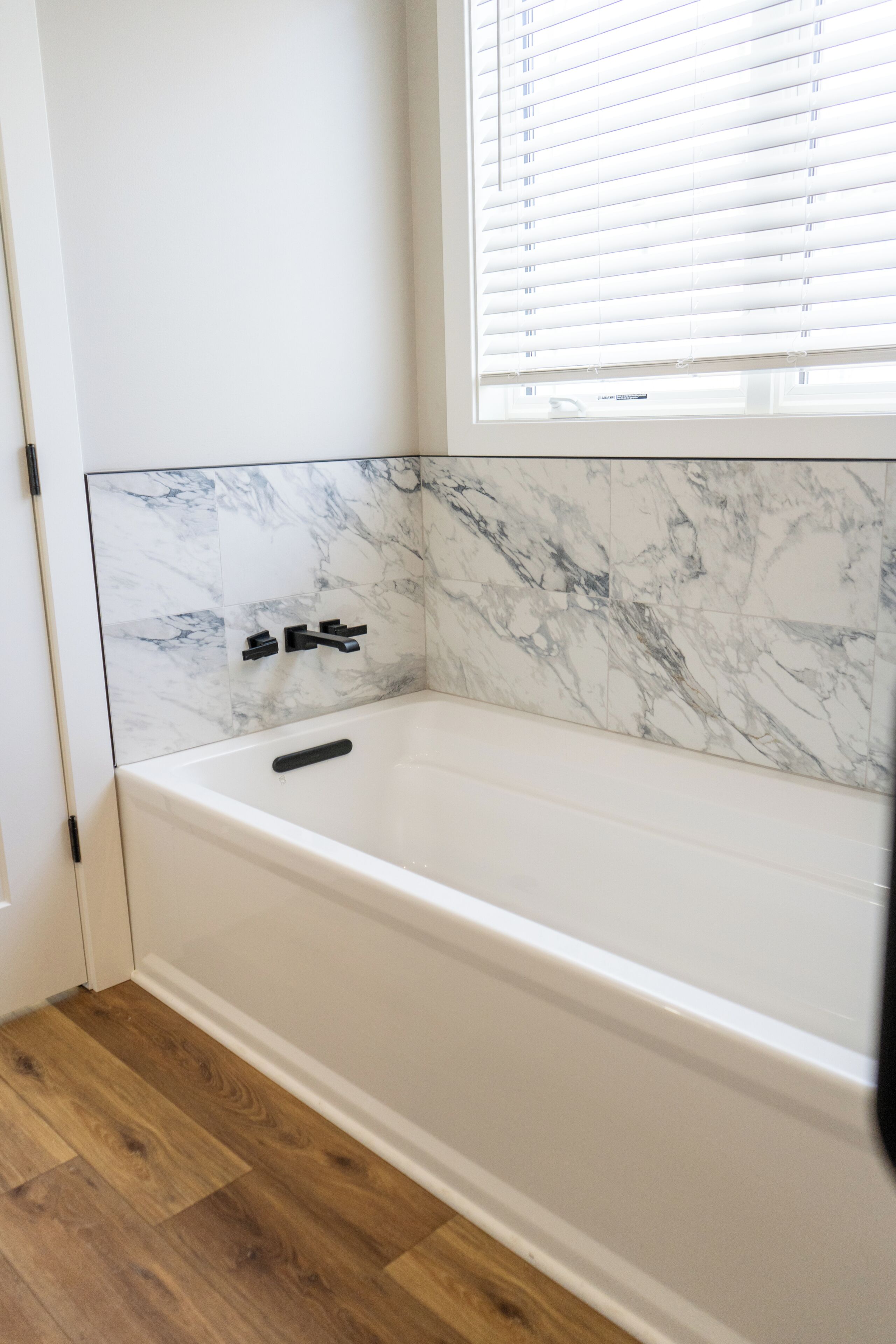 A bathroom with a white bathtub, marble wall tiles, and a window with blinds.