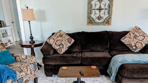 Ein Wohnzimmer mit braunem Sofa, einem gemusterten Sessel und Hocker, einem Couchtisch und einem Teppich mit Muster.