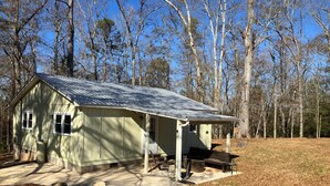 Eine kleine, eingeschossige Holzhütte mit Veranda und Bank, umgeben von Bäumen und blauem Himmel.