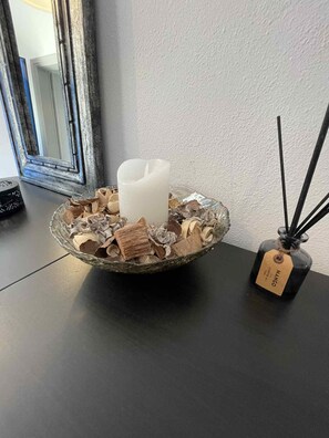 A candle in a glass bowl on a table, next to a black bottle with reed sticks. - Helios Studio in paradise Sisi (sisi)