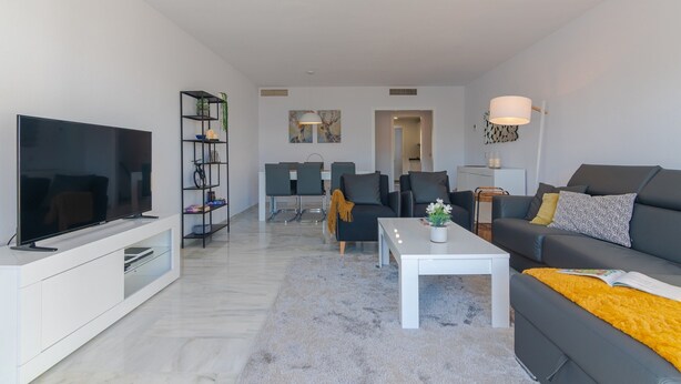 A modern living room with a flat-screen TV, a bookshelf, a sofa, a coffee table, and a dining area in the background. - Beachside Penthouse With Rooftop Terrace (Estepona)