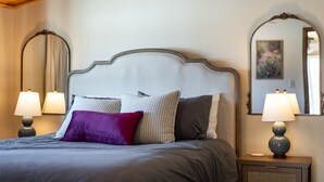 Una cama bien hecha con un cabecero gris, varios almohadones y dos lámparas de noche sobre mesitas de madera.