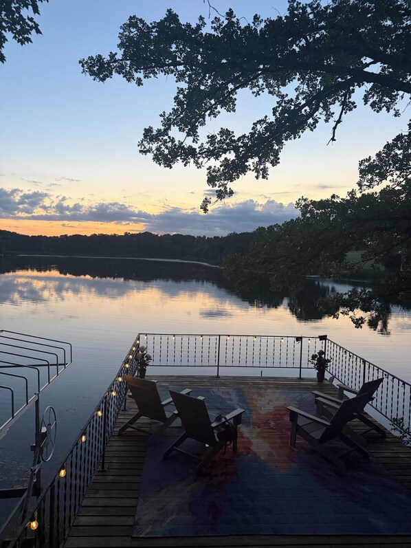 Eine Terrasse mit Stühlen und Geländer mit Blick auf einen stillen See bei Sonnenuntergang.