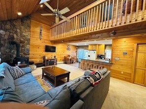 A log cabin interior with a stone fireplace, a wooden deck, a kitchen, and a living area with a sofa and coffee table.
